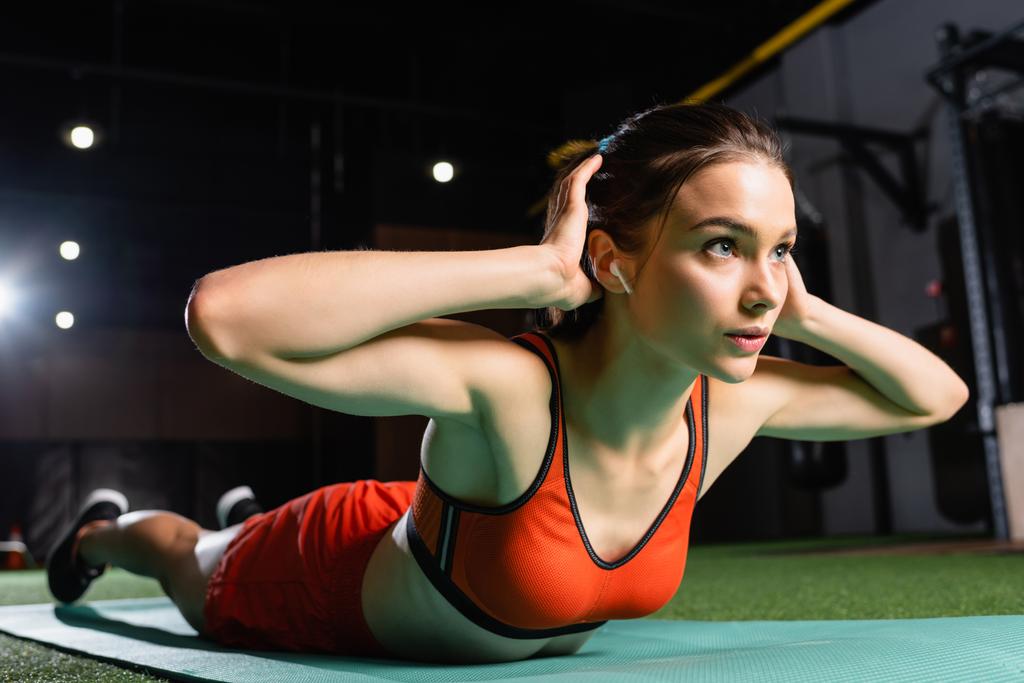 young sportswoman with earphone exercising on fitness mat in gym, blurred foreground - Photo, Image
