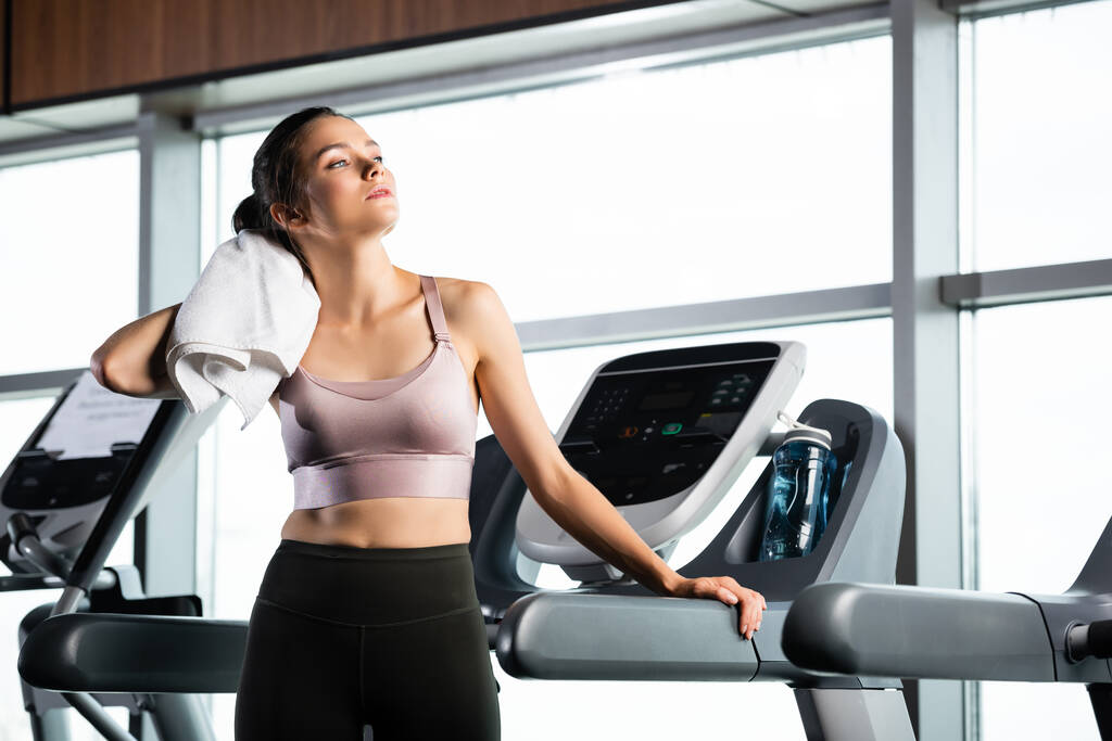 young sportswoman wiping neck with towel while standing on treadmill  - Photo, Image