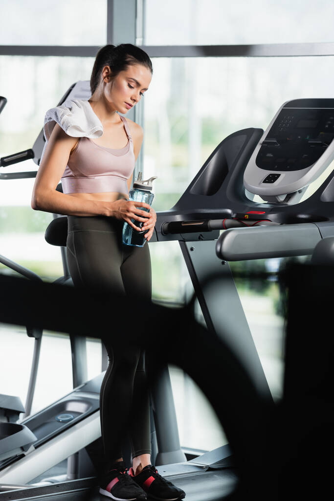 tired sportswoman standing on treadmill with closed eyes and holding sports bottle on blurred foreground - Photo, Image