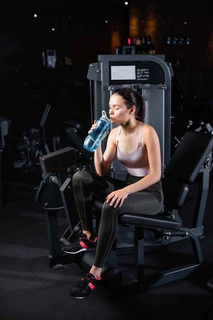 mladá sportovkyně sedící na tréninkovém stroji se zavřenýma očima a pijící ze sportovní láhve - Fotografie, Obrázek
