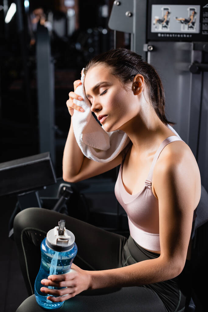 exhausted sportswoman wiping face with towel while sitting on training machine and holding sports bottle - Photo, Image