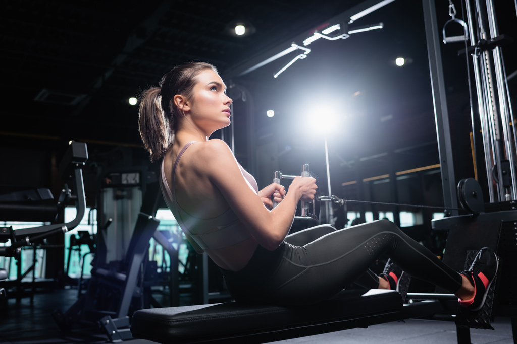 joven deportista mirando hacia otro lado mientras hace ejercicio en la máquina de remo en el gimnasio - Foto, Imagen