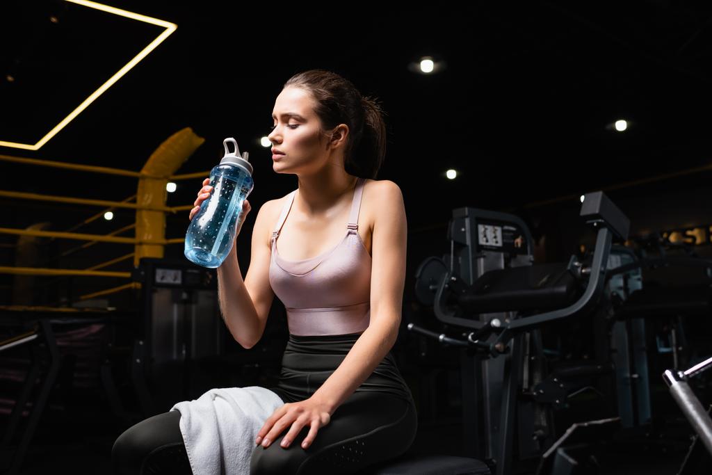 Yorgun sporcu kadın antrenman makinesinde gözleri kapalı ve elinde spor şişesiyle oturuyor. - Fotoğraf, Görsel
