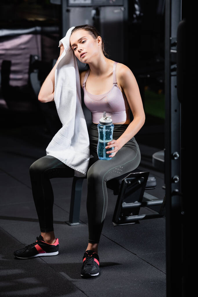 Müde Sportlerin wischt Kopf mit Handtuch ab, während sie mit Sportflasche auf Trainingsgerät sitzt - Foto, Bild