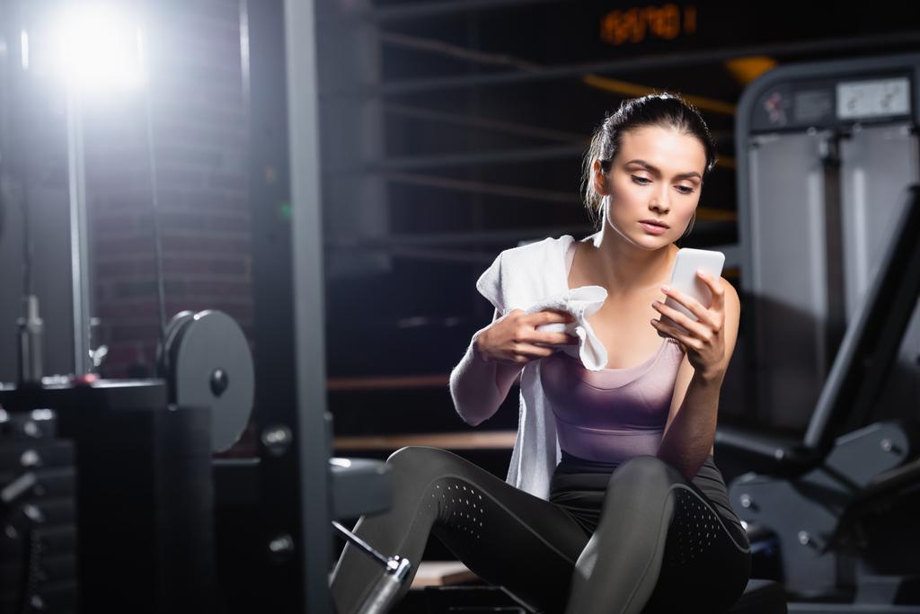 sportovkyně sedící na tréninkovém stroji a používající smartphone v posilovně na rozmazaném popředí - Fotografie, Obrázek
