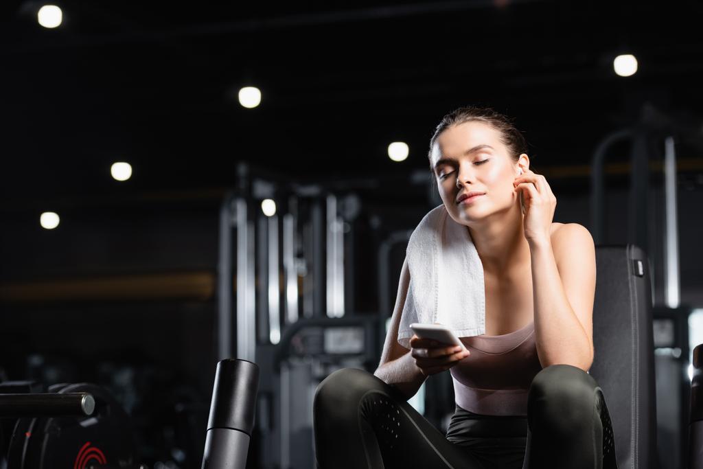 young sportswoman listening music in wireless earphone with closed eyes while holding smartphone in gym - Photo, Image