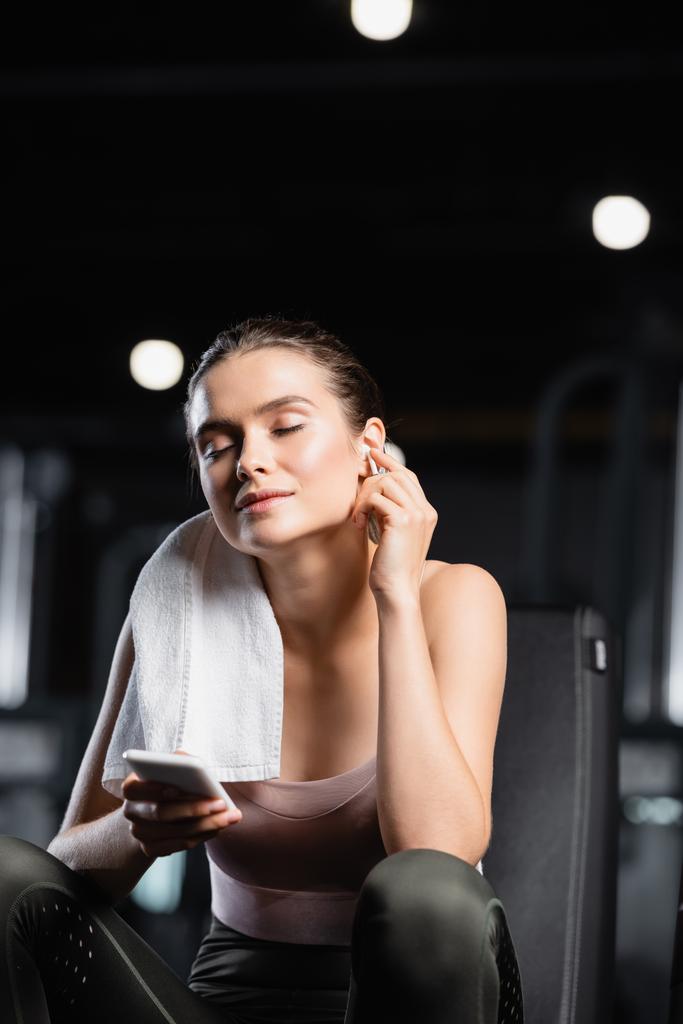 smiling sportswoman holding mobile phone and listening music in wireless earphone in sports center - Photo, Image