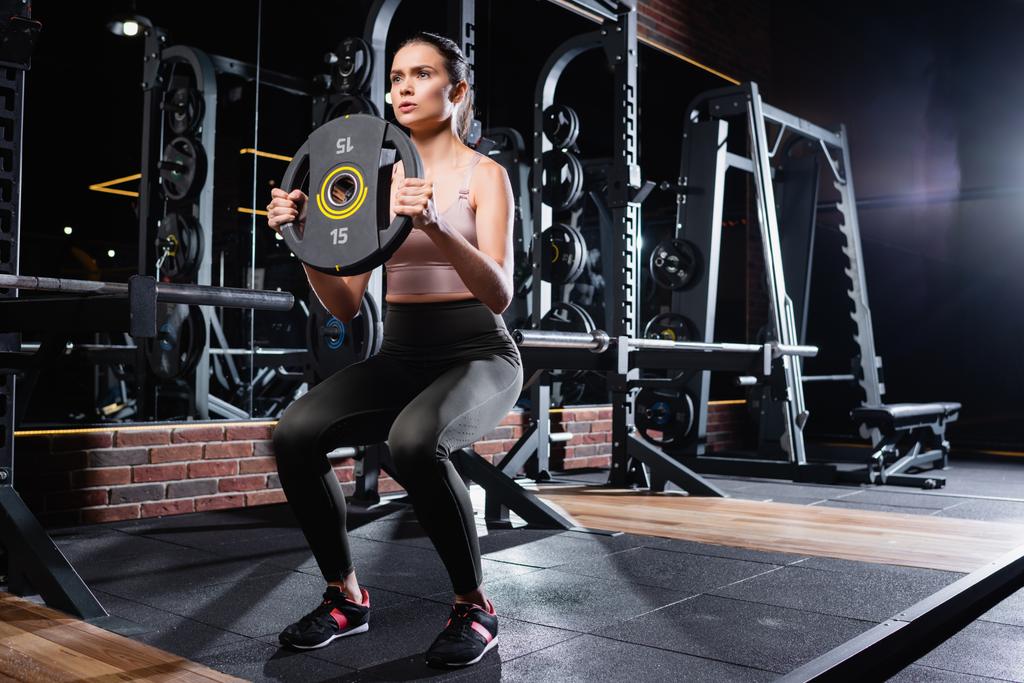 junge Sportlerin in Leggings und Turnschuhen beim Sit-Ups mit Gewichtsscheibe im Fitnessstudio - Foto, Bild
