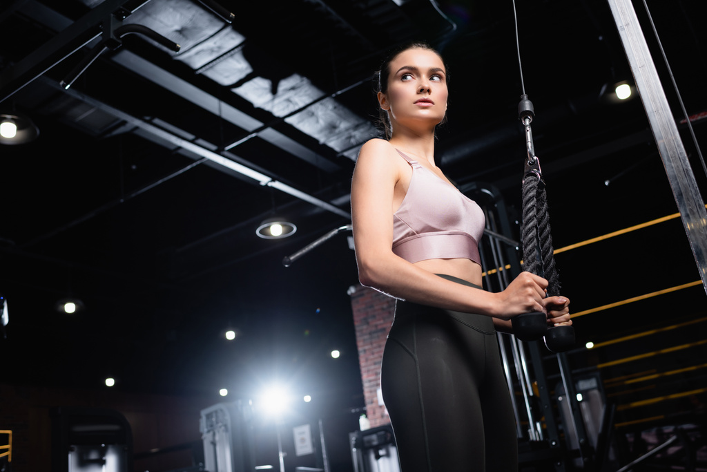 low angle view of sportswoman looking away while training on cable pushdown machine - Photo, Image