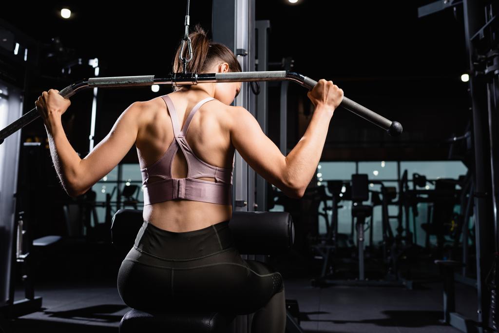 Вид сзади атлетичной женщины, делающей упражнения на растяжку спины и рук на лат-тренажере - Фото, изображение