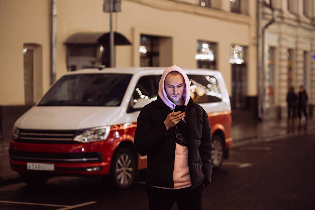 Man in hood looking into his smartphone in background of car on night street - Photo, Image