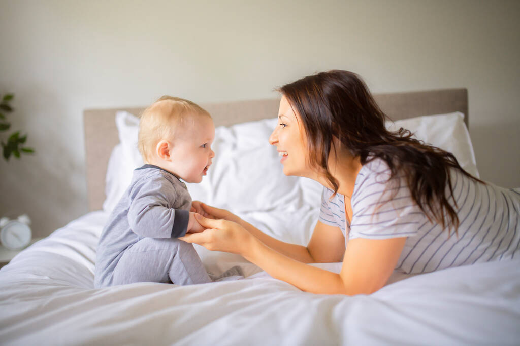 Mãe e criança em uma cama branca brincando no quarto ensolarado - Foto, Imagem