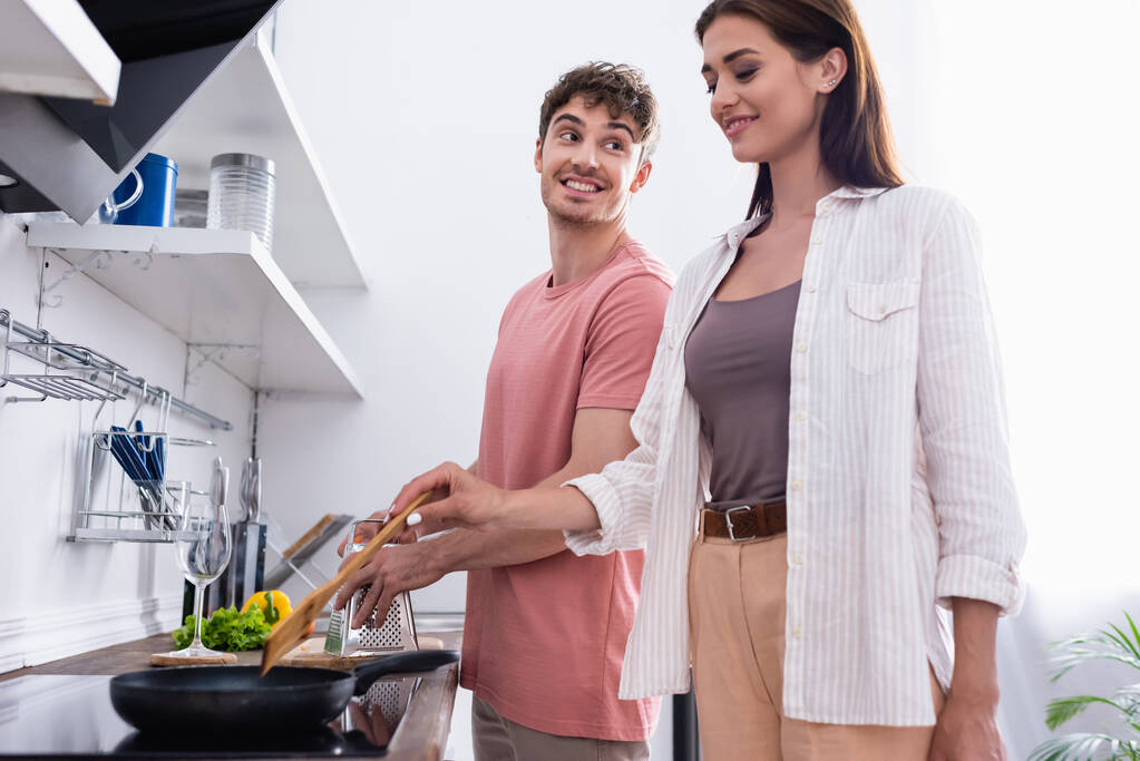 Усміхнений чоловік дивиться на дівчину, що готує на плиті на кухні
  - Фото, зображення