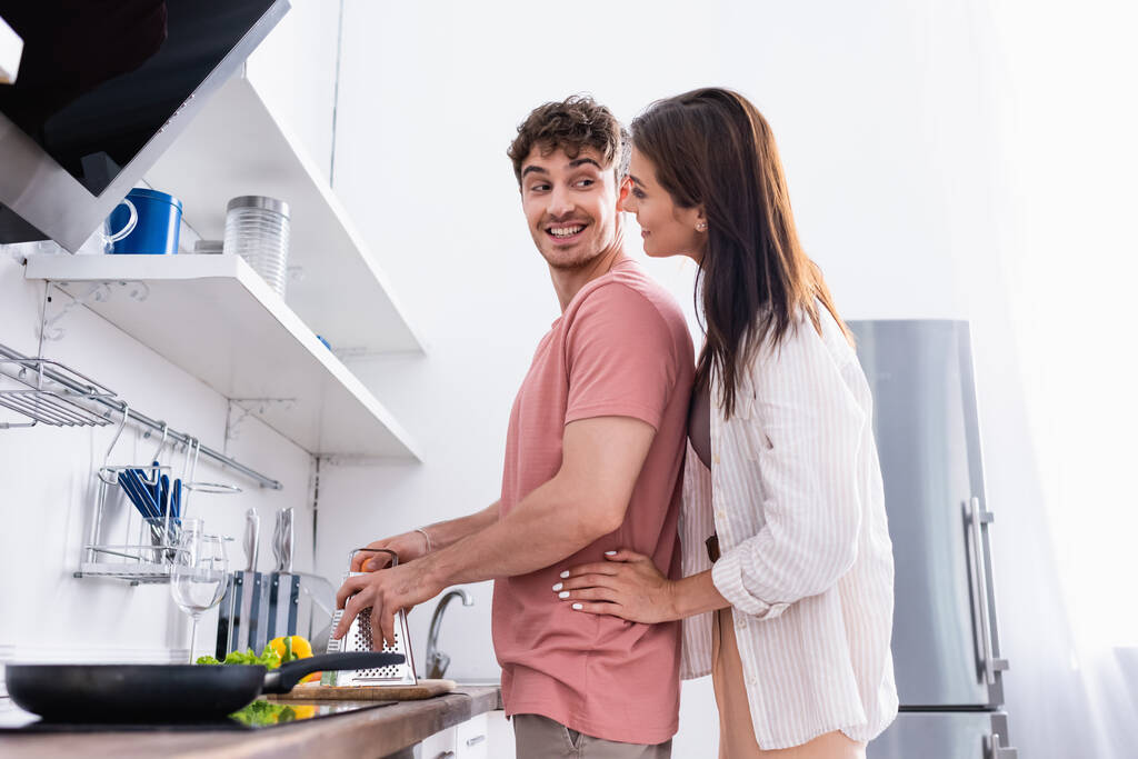 Widok z boku uśmiechnięta kobieta przytula chłopaka trzymając tarkę w pobliżu warzyw i patelni na kuchence na zamazanym pierwszym planie  - Zdjęcie, obraz