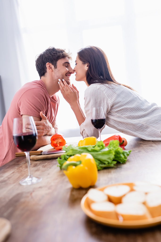 Mujer sonriente besando novio cerca de verduras y copas de vino en primer plano borrosa  - Foto, imagen