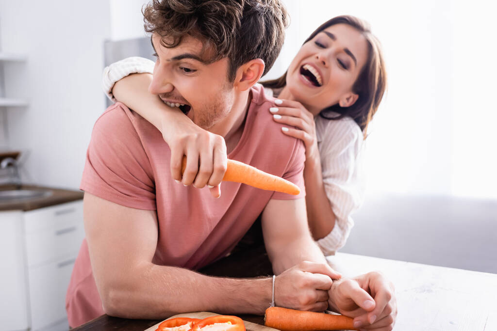 Мужчина кусает руку веселой девушки, держащей морковку на кухне.  - Фото, изображение