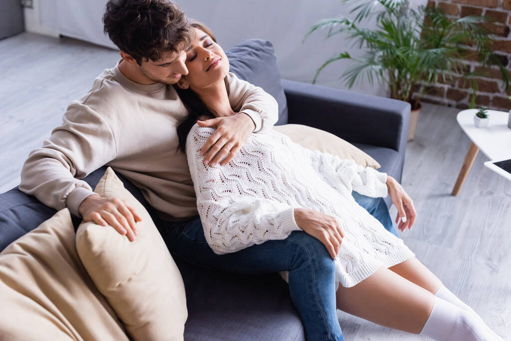 Молодий чоловік обіймає усміхнену жінку в светрі на дивані з подушками
  - Фото, зображення