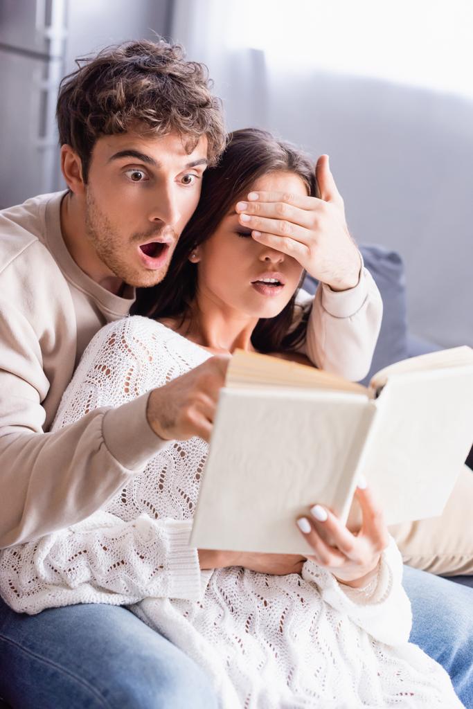 Homme excité couvrant les yeux de petite amie tout en lisant le livre sur le premier plan flou à la maison  - Photo, image