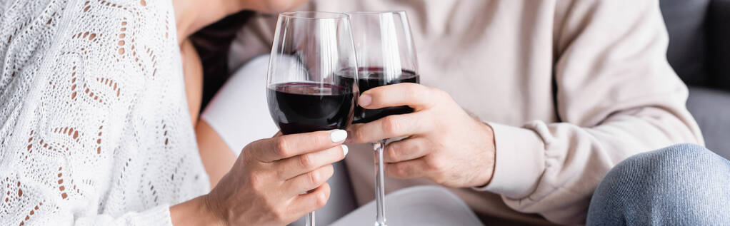 Vista recortada de copas de vino tinto en manos de parejas jóvenes sobre fondo borroso, pancarta  - Foto, imagen