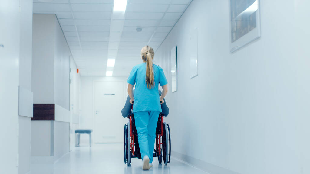Foto dell'infermiera che si muove paziente sulla sedia a rotelle attraverso il corridoio dell'ospedale. Procedura da seguire. Ospedale moderno luminoso con personale amichevole. - Foto, immagini