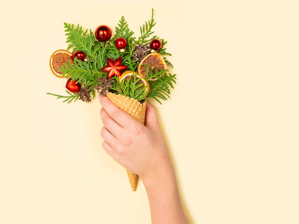 A female hand holds a waffle cone with thuja sprigs and Christmas toys on a yellow background. New Year card. Copy space, flat lay. The concept photo is an original Christmas present. - Photo, Image