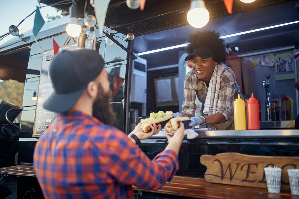 educada afro-americana empregada segurando, dando, duas sanduíches para um cliente caucasiano barbudo satisfeito. serviço de fast food - Foto, Imagem