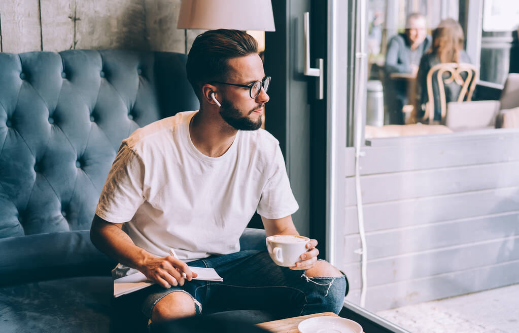 Homem pensativo em roupas casuais e óculos escrevendo poesia apreciando música através de fones de ouvido sem fio e bebendo café enquanto sentado no sofá no café e olhando para fora da janela - Foto, Imagem