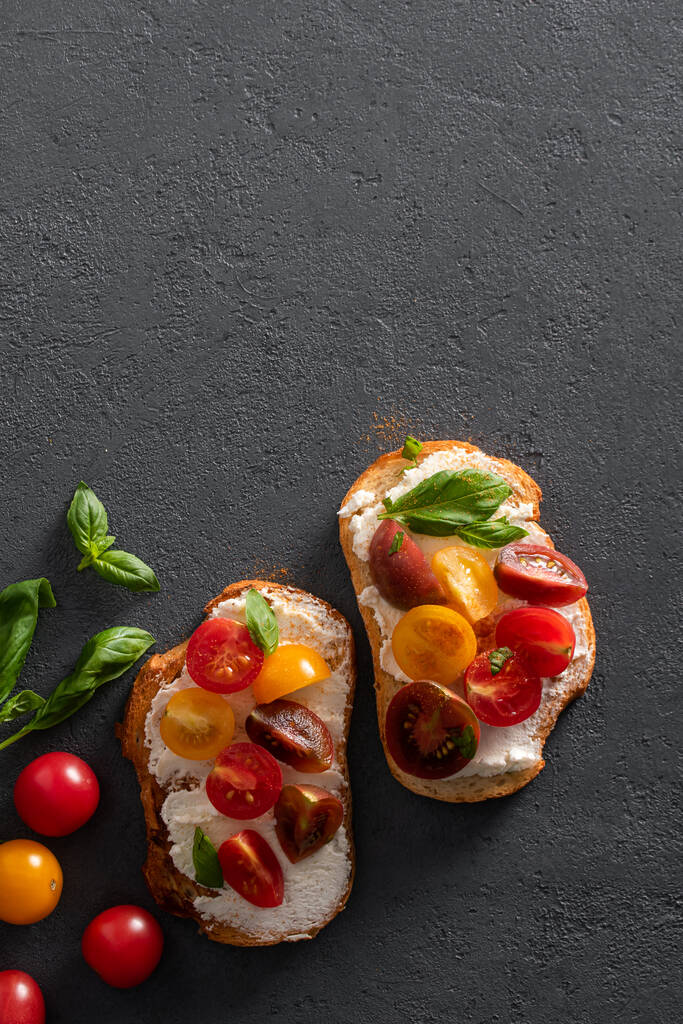 Poser à plat avec deux bruschetta au fromage caillé, tomates cerises et basilic sur un fond de béton foncé. Cuisine italienne, sandwichs au petit déjeuner. - Photo, image