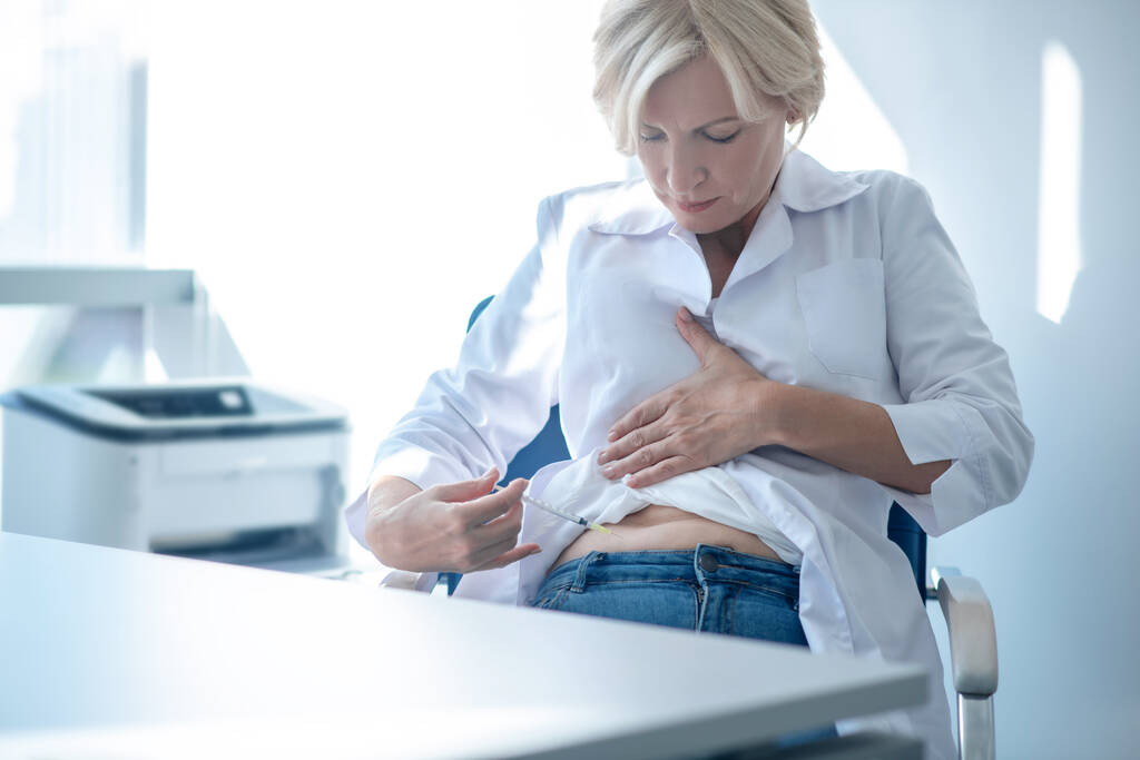 Reife blonde Frau sitzt am Tisch und spritzt Insulin in den Bauch - Foto, Bild