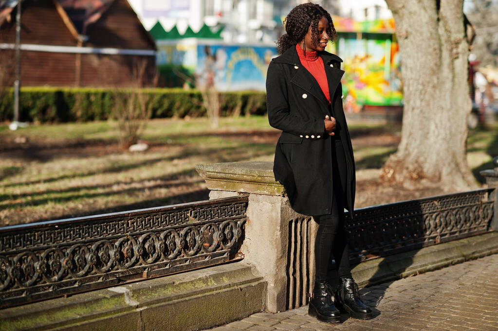 Portré egy göndör hajú afrikai nő visel divatos fekete kabát és piros garbó pózol szabadban napsütéses napon. - Fotó, kép