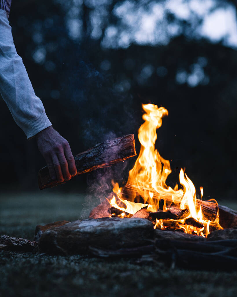 Ein Mann legt abends einen Baumstamm in ein Lagerfeuer - Foto, Bild