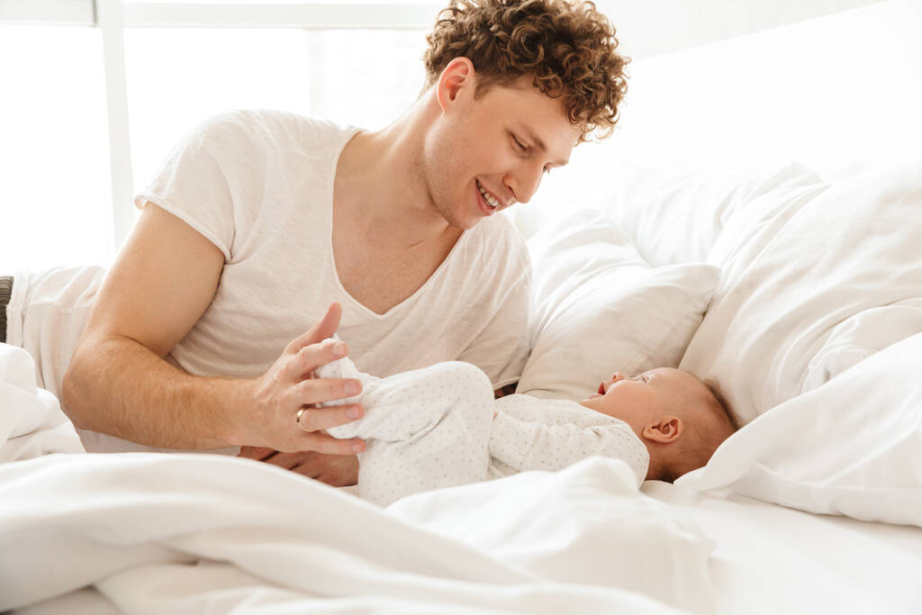 Счастливый молодой отец, играя со своим маленьким сынишкой, лежа в постели дома, обнимая - Фото, изображение