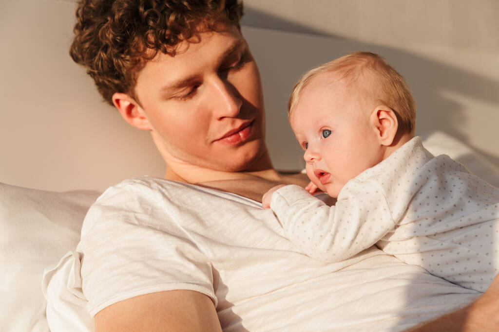 幸せな若いです父と遊ぶ彼の小さなかわいい赤ちゃんの息子と一緒にベッドで家に横たわって、抱擁 - 写真・画像