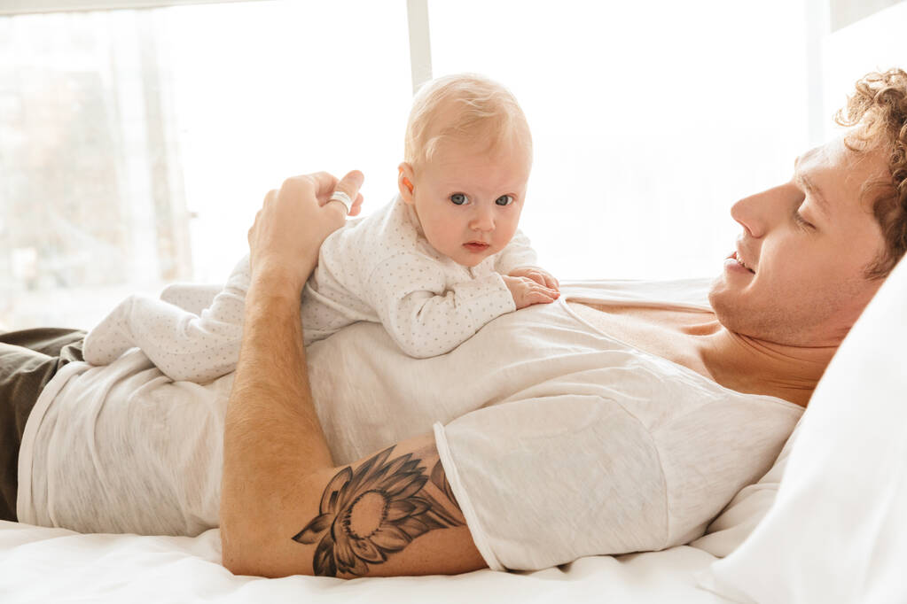 gelukkig jong vader spelen met zijn kleine cutr baby zoon terwijl liggend in bed thuis, omarmen - Foto, afbeelding