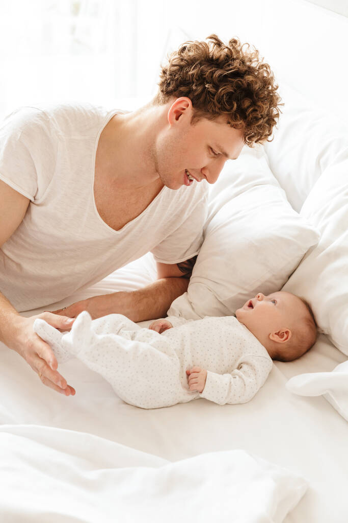 幸せな若いです父と遊ぶ彼の小さなかわいい赤ちゃんの息子と一緒にベッドで家に横たわって、抱擁 - 写真・画像