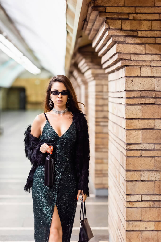 mulher elegante em vestido lurex preto de pé com garrafa de vinho e bolsa na estação subterrânea - Foto, Imagem