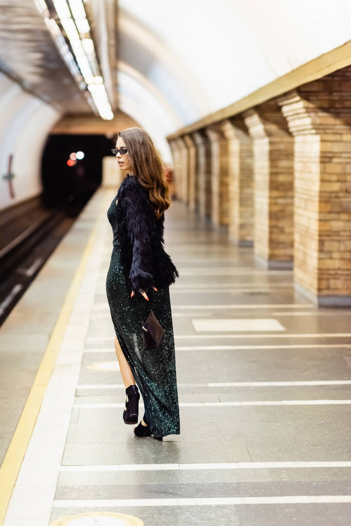 κομψή γυναίκα με μαύρο φόρεμα lurex και γυαλιά ηλίου περπατώντας κατά μήκος της πλατφόρμας του μετρό - Φωτογραφία, εικόνα