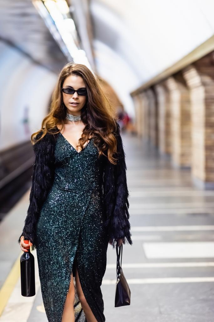 Metroda şarap şişesi tutan siyah lurex elbiseli ve güneş gözlüklü baştan çıkarıcı kadın. - Fotoğraf, Görsel