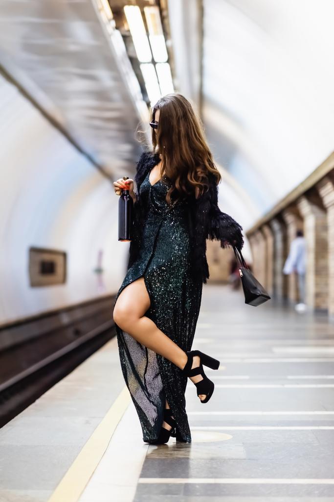 mulher glamour em lurex vestido preto segurando garrafa de vinho enquanto posando na plataforma do metrô - Foto, Imagem