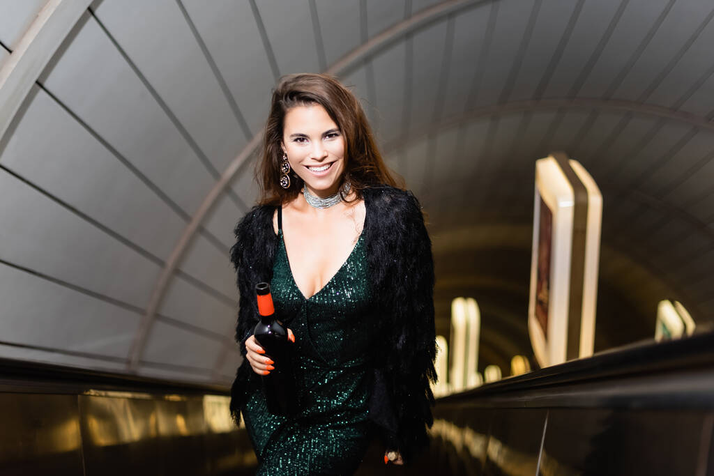 μοντέρνα γυναίκα σε μαύρο φόρεμα χαμογελώντας στην κάμερα, ενώ κρατώντας μπουκάλι κρασί στις κυλιόμενες σκάλες - Φωτογραφία, εικόνα