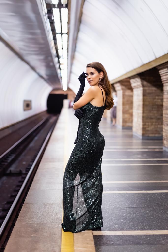 donna sensuale in abito lungo nero guardando la fotocamera mentre in piedi sulla piattaforma sotterranea - Foto, immagini