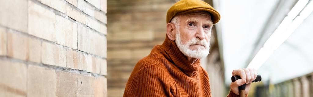 senior man in sweater and cap looking away while sitting on metro platform on blurred foreground, banner - Photo, Image