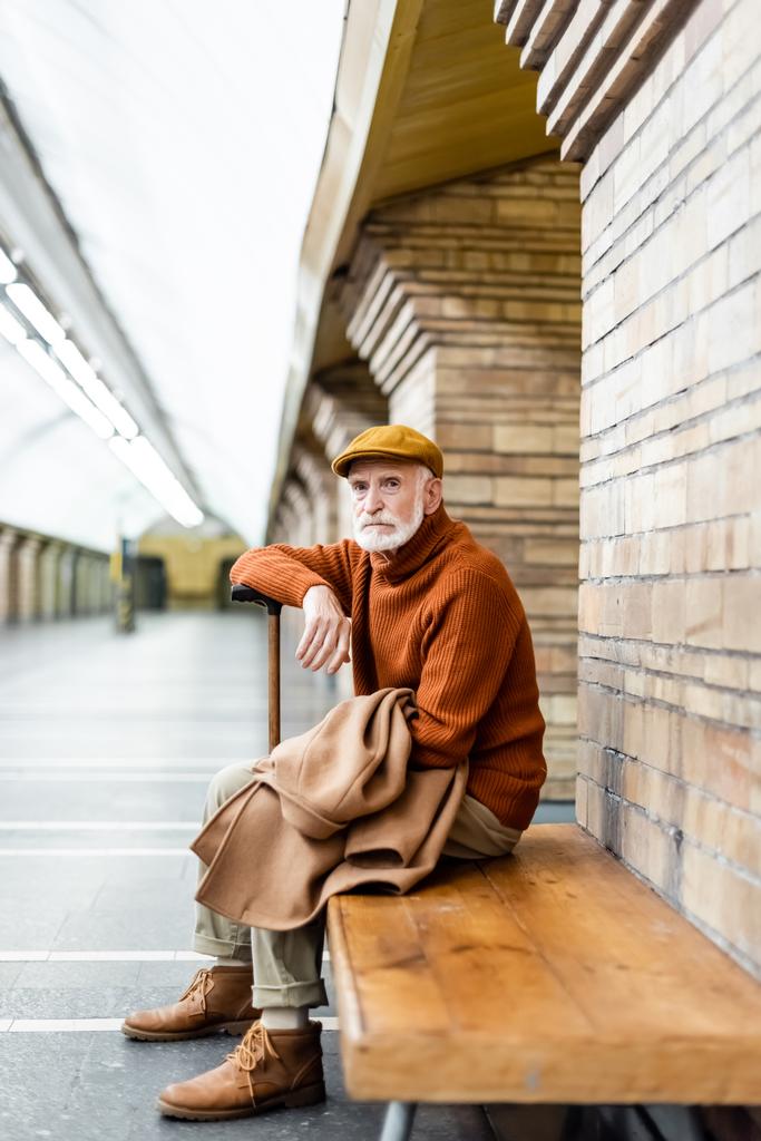 ηλικιωμένος άνδρας σε φθινοπωρινή στολή ακουμπά στο μπαστούνι, ενώ κάθεται στο παγκάκι πλατφόρμα του μετρό - Φωτογραφία, εικόνα