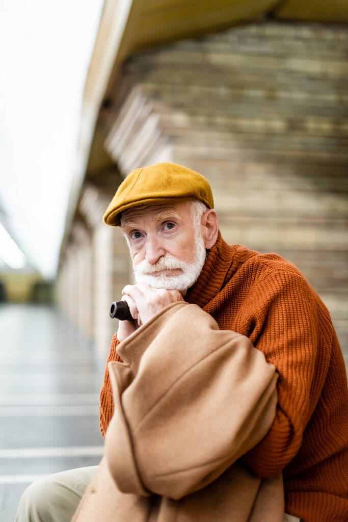 idős férfi sapkában és pulóverben néz félre, miközben ül sétapálcával a metró peronján - Fotó, kép