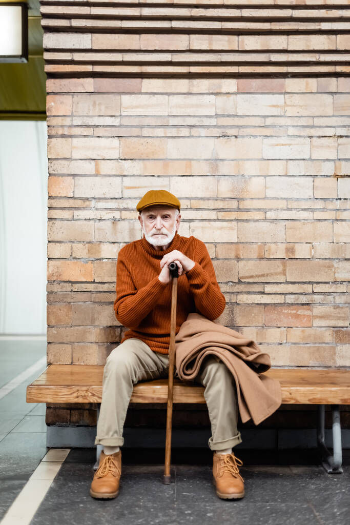 idős férfi őszi ruhában ül sétapálcával a metró peron padján - Fotó, kép