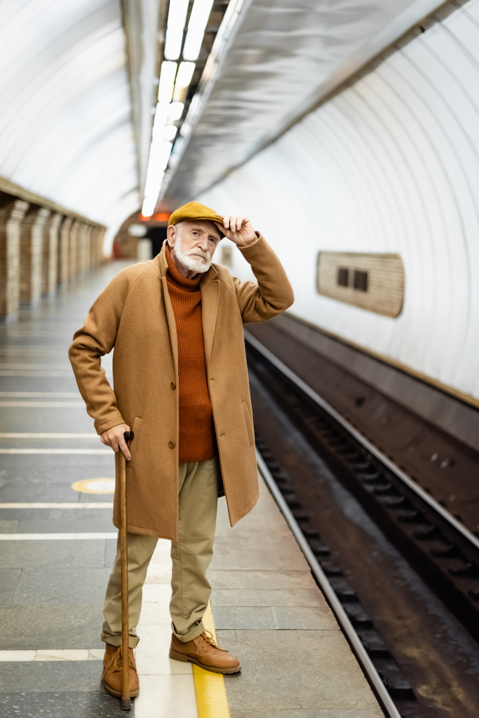 senior man in autumn outfit touching cap while standing on metro station platform - Foto, Imagen