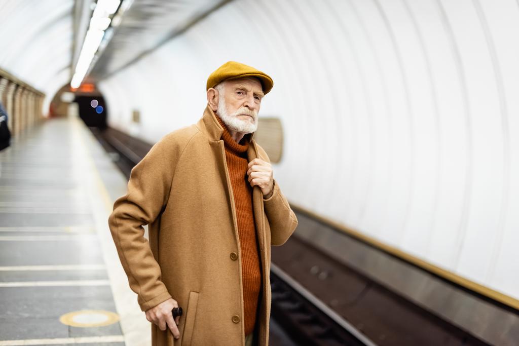 старший мужчина в осеннем наряде трогает воротник пальто, глядя на платформу метро - Фото, изображение