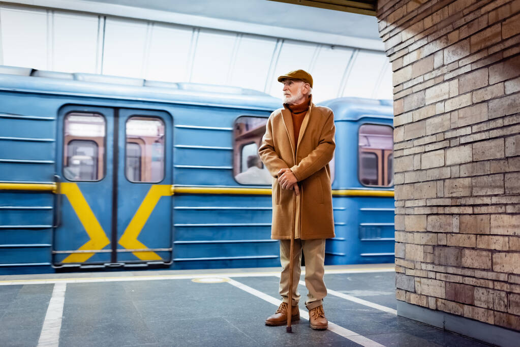 秋の服装の老人が電車のそばの地下鉄のホームで杖を持って立っていて - 写真・画像