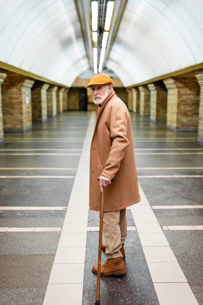 aged man in autumn coat and cap standing with walking stick at metro station - Foto, imagen