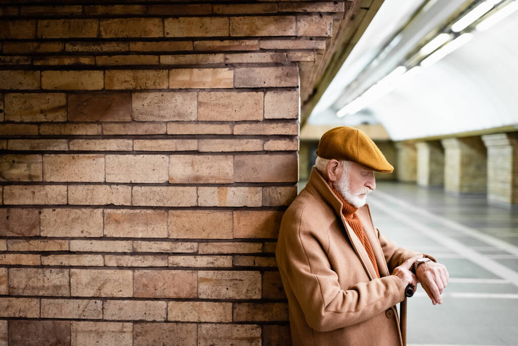 senior man in autumn outfit looking at wristwatch while leaning on column at metro station - Photo, Image